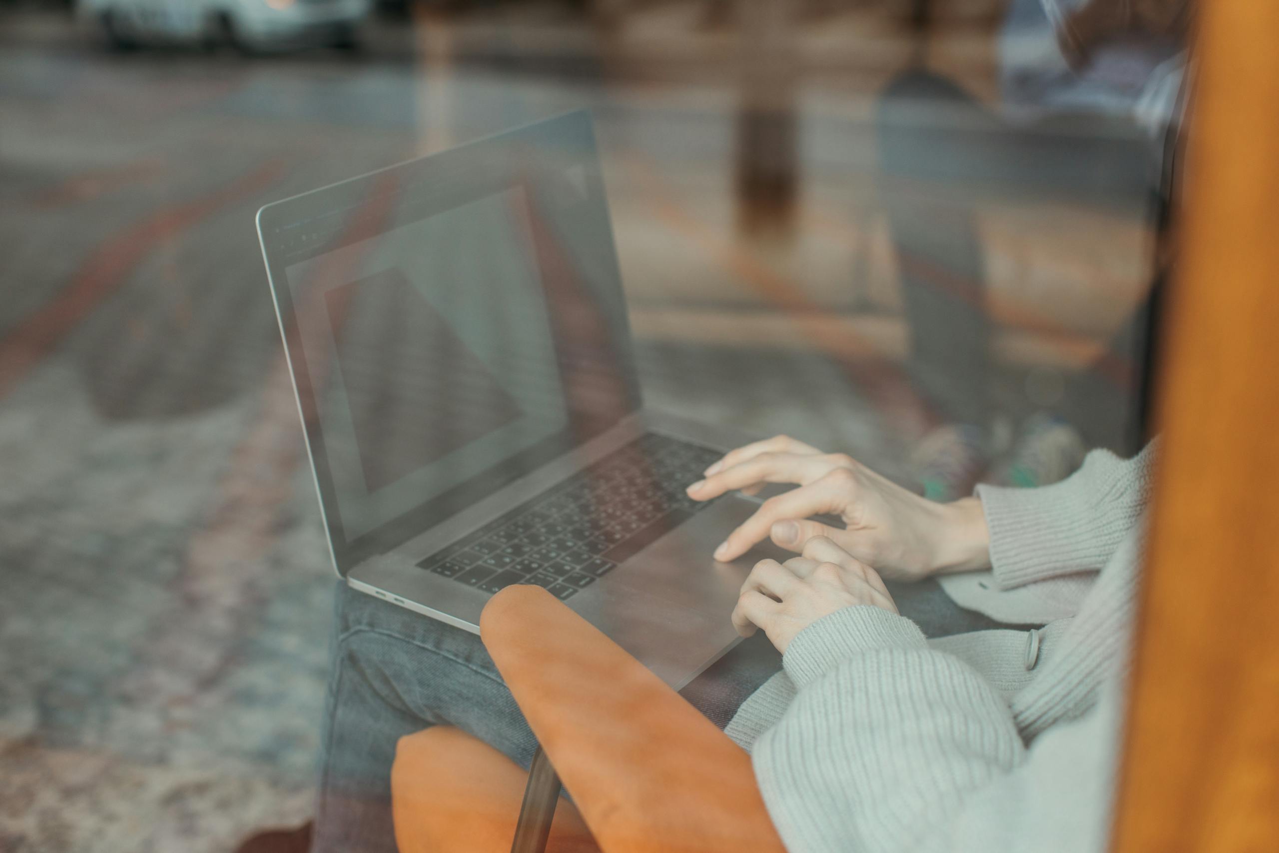 A person types on a laptop indoors, reflected through a window, wearing a comfy sweater.