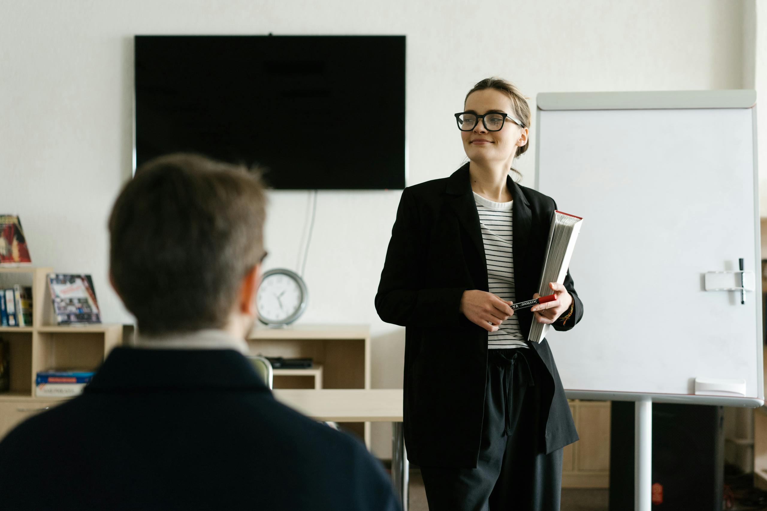 Caucasian female teacher in black blazer holding books, presenting to a student in a classroom.